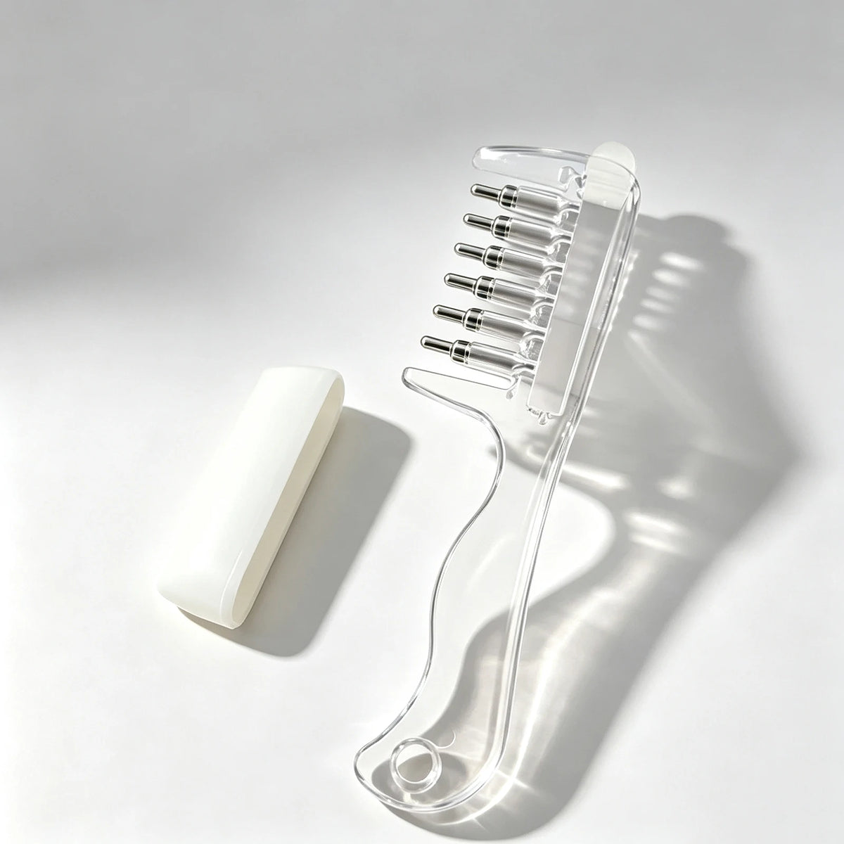 Clear refillable scalp massage comb with roller ball applicator resting on a white surface.