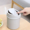 A hand is flipping the lid of a compact, light gray desktop trash bin.