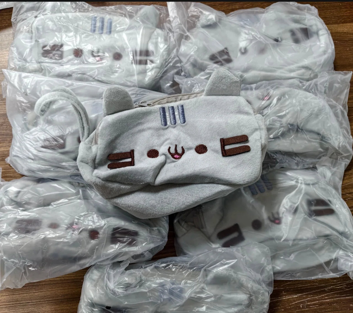 A pile of cute cat plush pencil cases with zipper closures in light gray fabric, packaged in clear plastic bags.