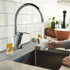 A hand holds a glass of water dripping from a modern kitchen faucet over a silicone drain mat.