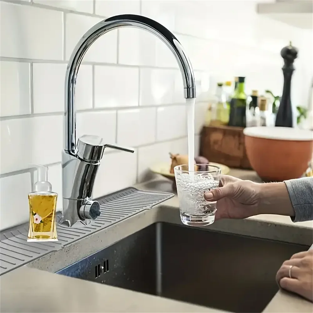 A hand holds a glass of water dripping from a modern kitchen faucet over a silicone drain mat.