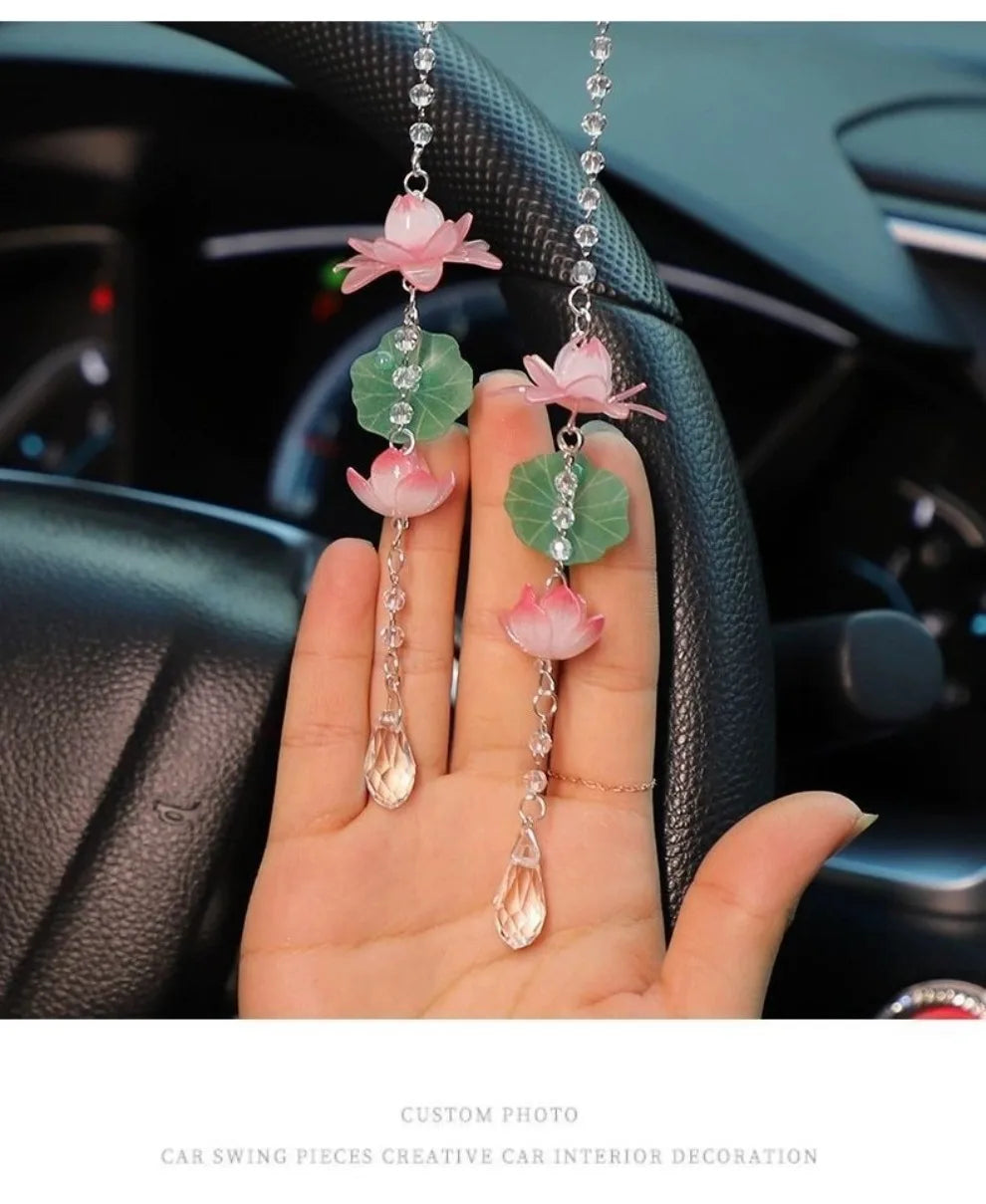 A hand holds several pink lotus car pendants featuring green leaves and clear beads against a dark car interior background.