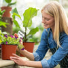 A blonde woman is potting a plant in an orange terracotta pot, surrounded by green foliage.