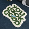 A white non-slip letter rug with the word GOOD you spelled out in green embroidery on a dark blue surface.