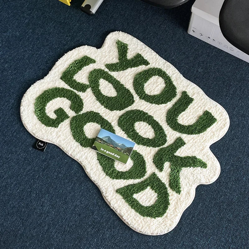 A white non-slip letter rug with the word GOOD you spelled out in green embroidery on a dark blue surface.