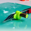 A bright green magnetic film cutting guide with a sharp blade is shown resting on a teal surface.