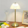 A brass-toned table lamp with a translucent dome shade sits on a wooden surface next to a wine glass and a book.
