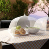 A collapsible mesh food cover is placed over a plate of pastries and a bowl on an outdoor table.