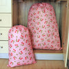 A pink drawstring laundry bag with a red and green cherry pattern is displayed in a wooden cabinet.