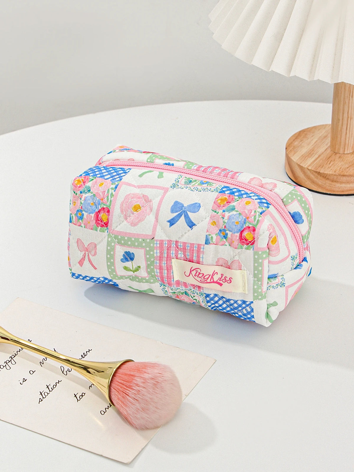 A pink and blue patchwork quilted makeup bag with floral embroidery rests on a white surface next to a makeup brush.