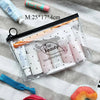 A clear plastic travel toiletry bag filled with various beauty products and a hanging hook on a striped cloth.