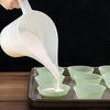 A hand pours white liquid from a plastic measuring cup with a long spout into small green paper cups.