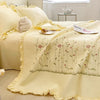 A pale yellow quilted bedspread with embroidered floral patterns and ruffled edges is displayed on a bed.