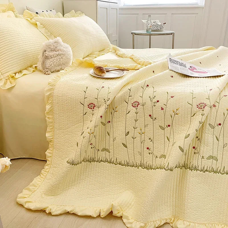 A pale yellow quilted bedspread with embroidered floral patterns and ruffled edges is displayed on a bed.