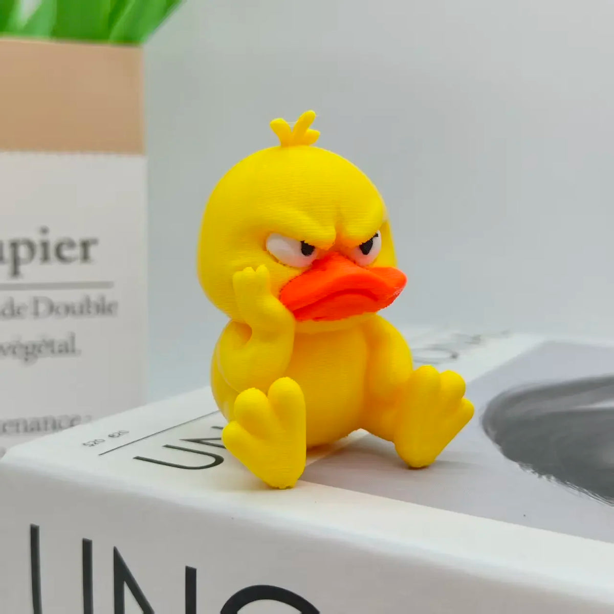 A bright yellow 3D printed duck statue with an angry expression sitting on a white box.
