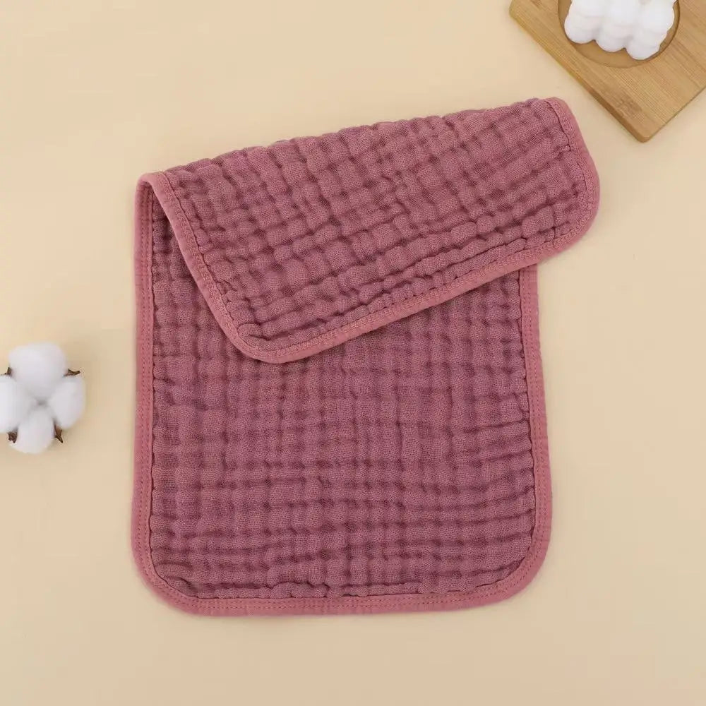 A stack of pink quilted muslin baby burp cloths with a matching cloth cover and a wooden spoon in the background.