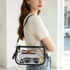 A woman holds a clear PVC shoulder bag with an adjustable strap and a brown leather accent against a bright background.