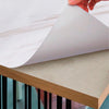 A hand is peeling a sheet of marble peel-and-stick wallpaper onto a wooden surface.