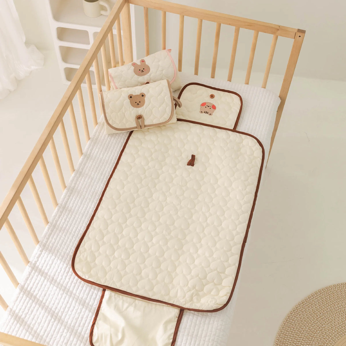A beige foldable portable diaper changing pad is placed inside a wooden crib with white bedding.