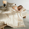 A woman rests on a bed covered with a luxurious champagne silk duvet and pillows.