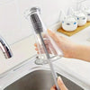 A hand holds a clear silicone bottle brush with a long handle, cleaning items in a kitchen sink.