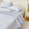 A light purple floral quilted bedspread with blue trim is displayed on a bed, next to a white headboard and pillows.