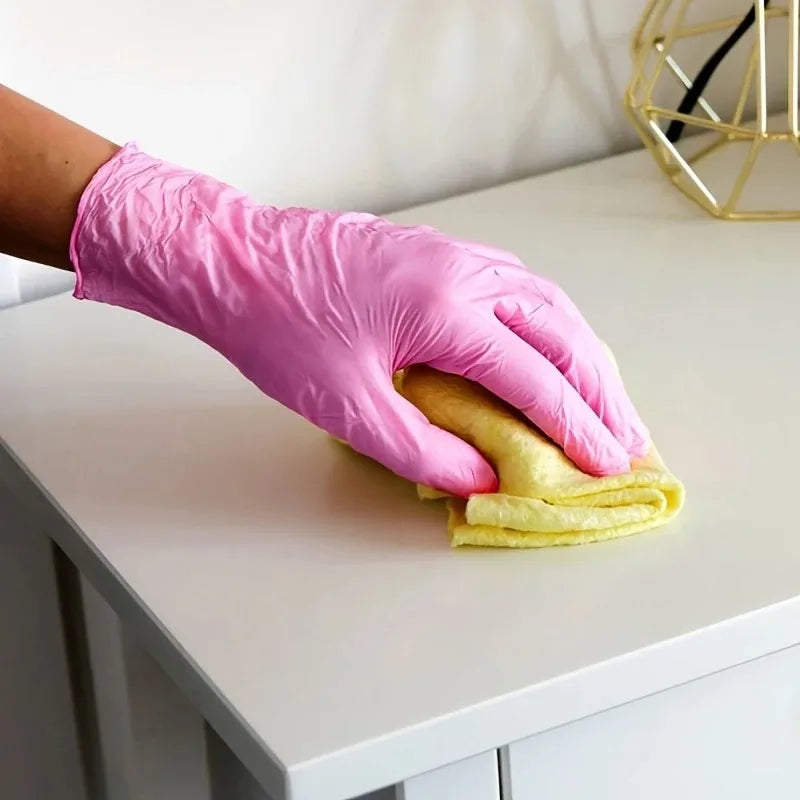 A hand wearing a pink disposable nitrile glove is holding a yellow cloth on a white surface.