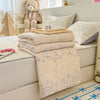 A beige lace-trim breathable summer duvet is folded on a white surface next to pillows and a teddy bear.