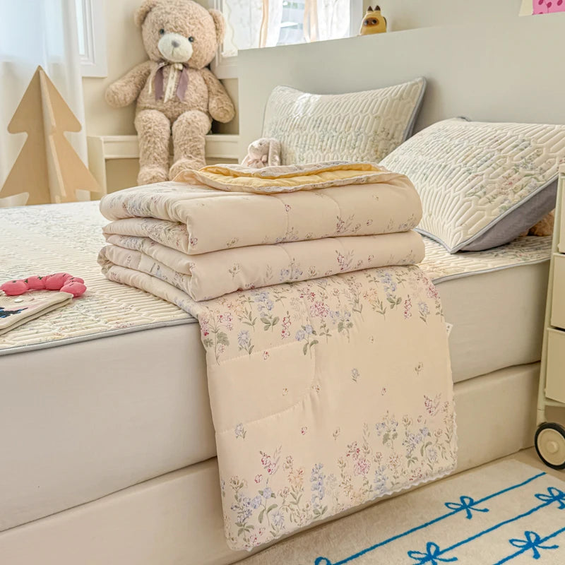 A beige lace-trim breathable summer duvet is folded on a white surface next to pillows and a teddy bear.