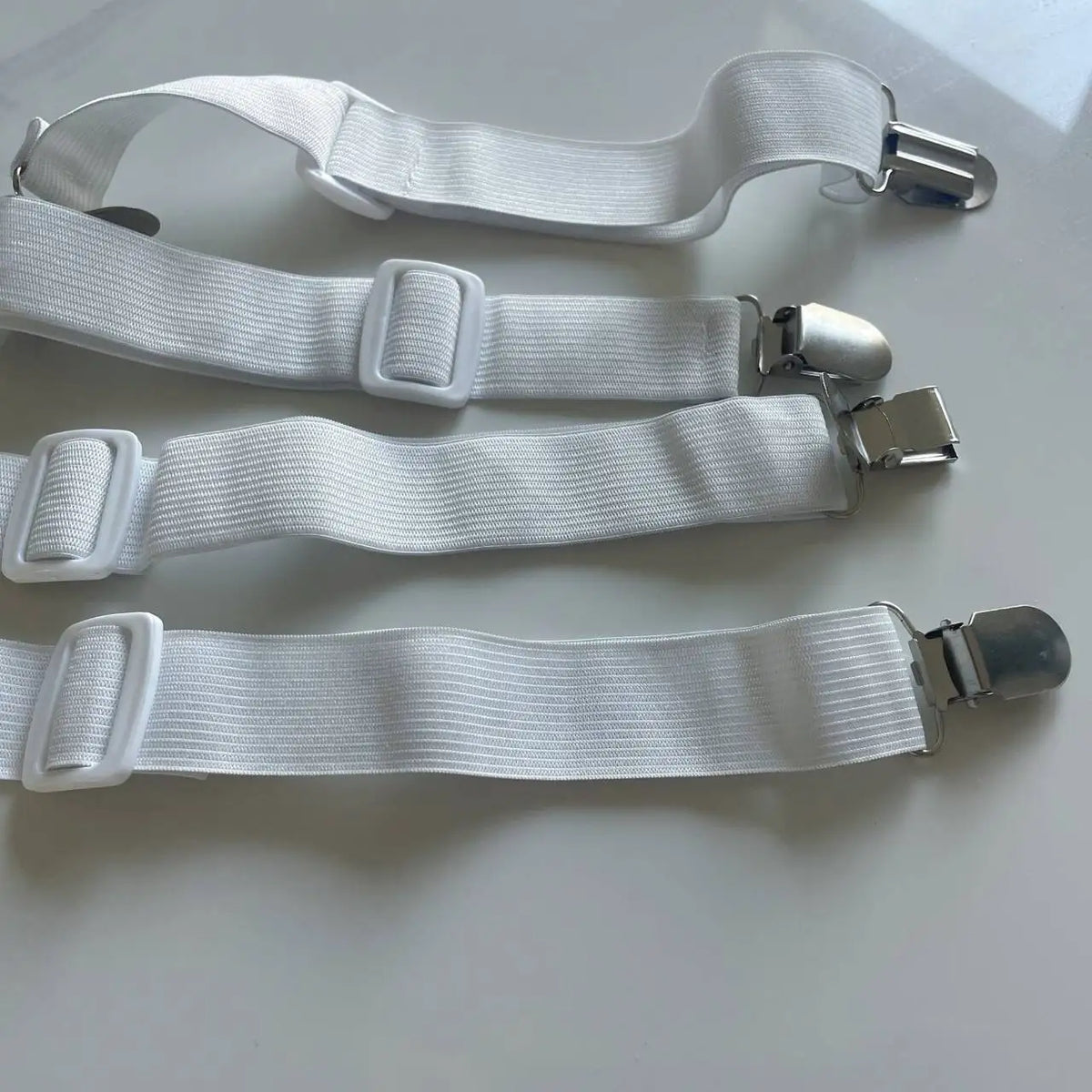 A pack of four white adjustable bed sheet grippers with strong clips laid out on a white surface.