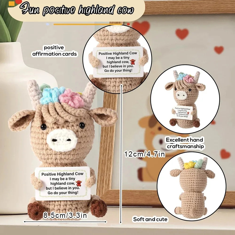 A handmade crochet Highland cow plush doll with a positive affirmation card and a wooden display stand.