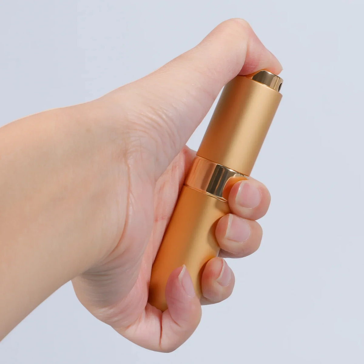 A hand holds a gold-colored refillable aluminum perfume atomizer for travel.