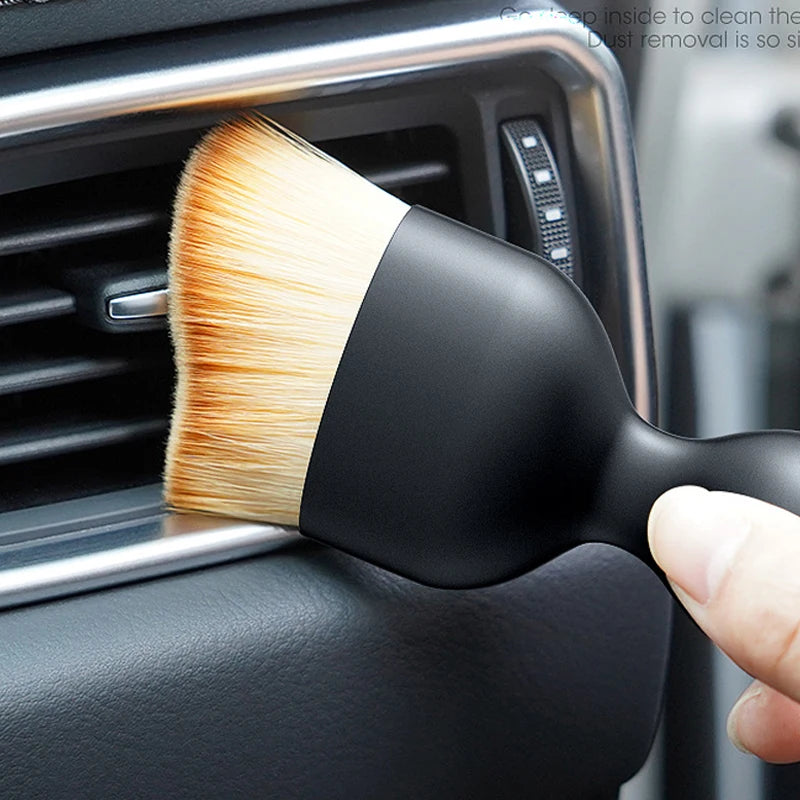 A black cleaning brush with soft, light-colored bristles is shown being used on a car vent or crevice.