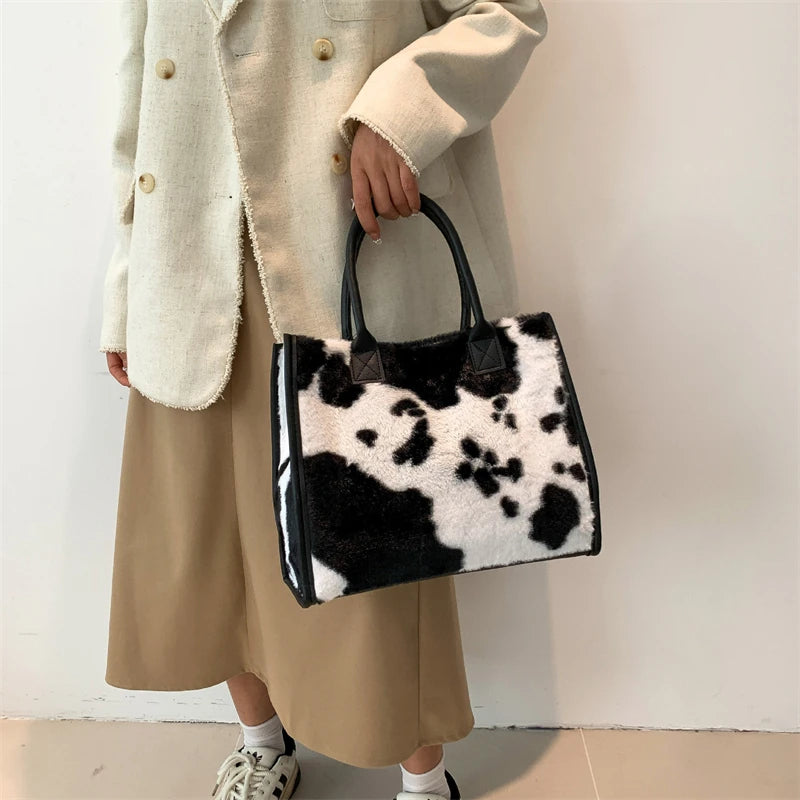 A woman holds a black and white faux fur leopard print tote bag while wearing a beige coat and skirt.