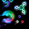 Hands manipulate glowing LED fidget spinners of various colors and shapes against a dark background.