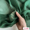 A hand is folding a piece of sheer, olive green mulberry silk chiffon fabric.
