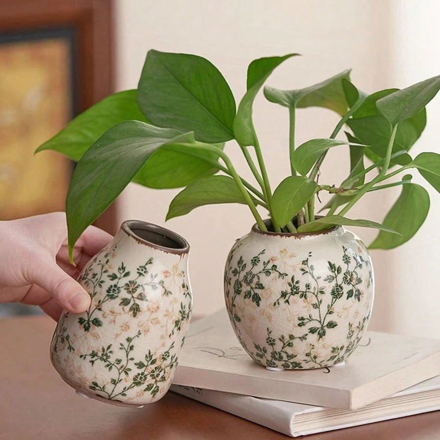 Eine Hand hält eine handgefertigte, gesplitterte Keramikvase mit grünen Blättern auf einer hellen Oberfläche.