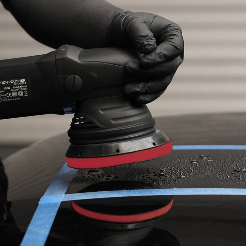 A hand wearing a black glove is using a paint polisher on a blue clay pad for car detailing.