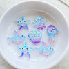 A white bowl contains various iridescent blue and purple resin charms shaped like starfish, fish, and a small purple creature