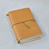 A tan leather refillable journal with a leather strap and a gold-colored metal tag resting on a white textured surface.