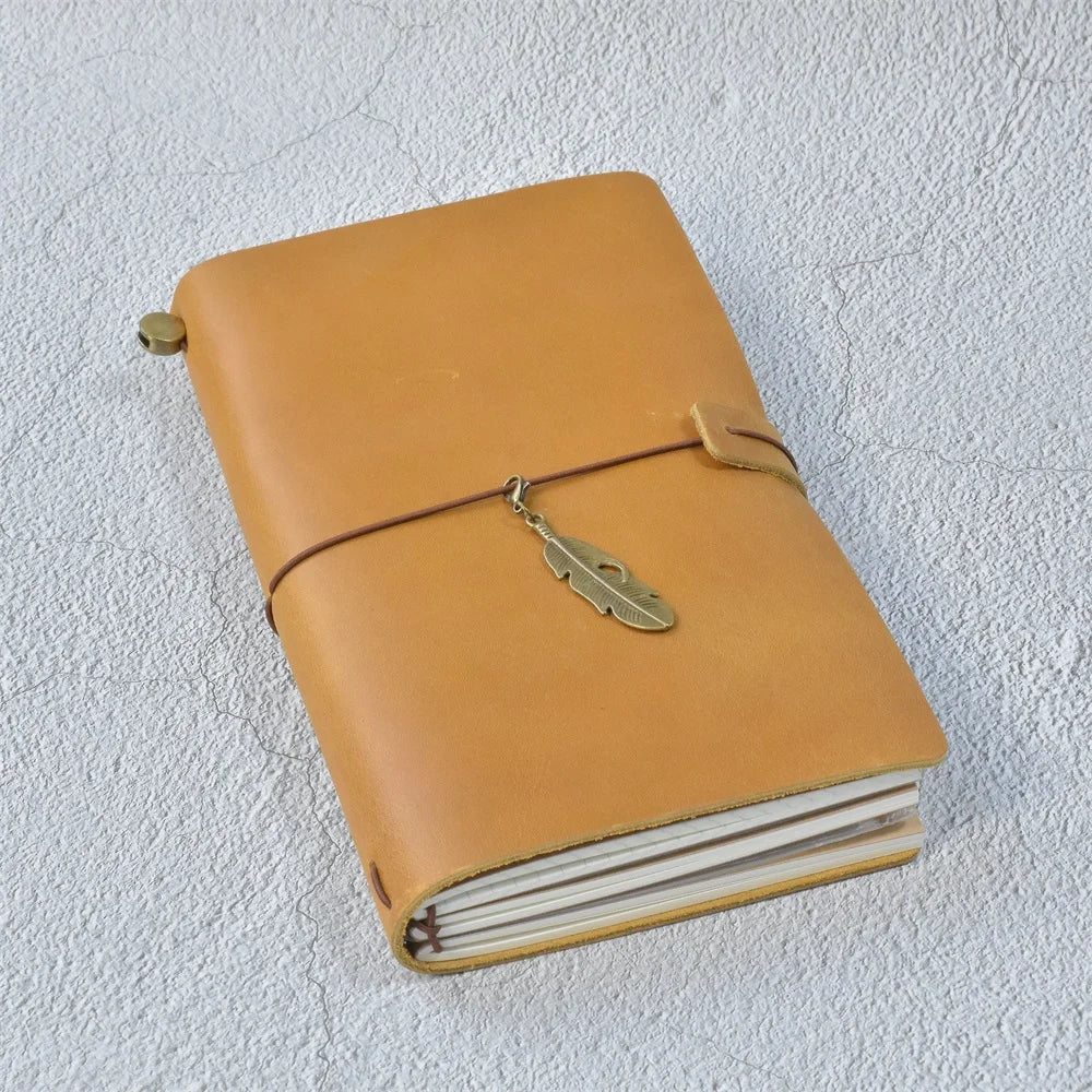 A tan leather refillable journal with a leather strap and a gold-colored metal tag resting on a white textured surface.