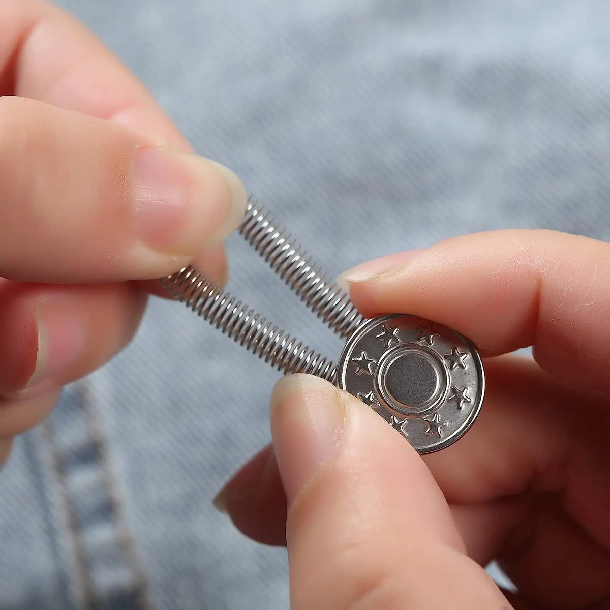 A hand is holding and manipulating an adjustable elastic waist extender button, which is a silver metal piece with a threaded