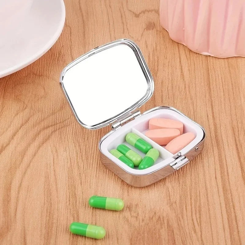 A portable metal pill organizer with a hinged lid is shown open, displaying several pink and green pills on a wooden surface.