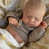 A newborn baby doll with soft skin and light brown hair is sleeping on a patterned fabric.