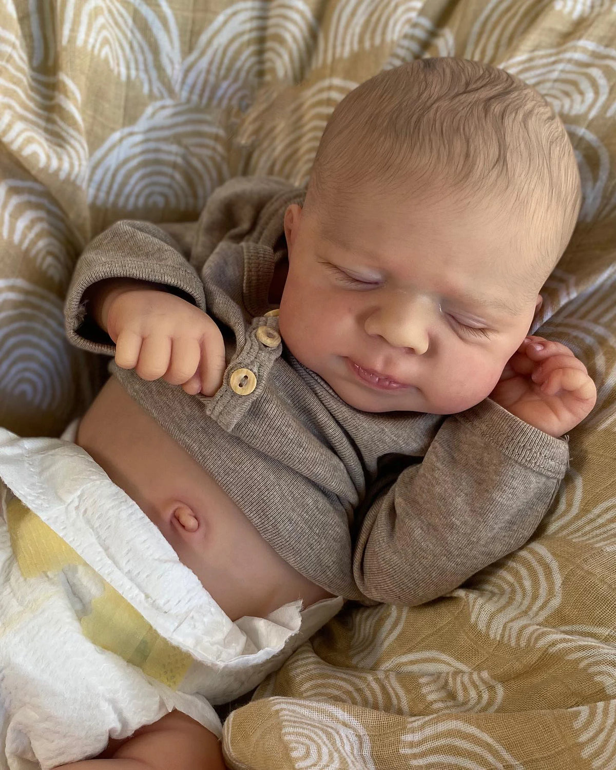 A newborn baby doll with soft skin and light brown hair is sleeping on a patterned fabric.