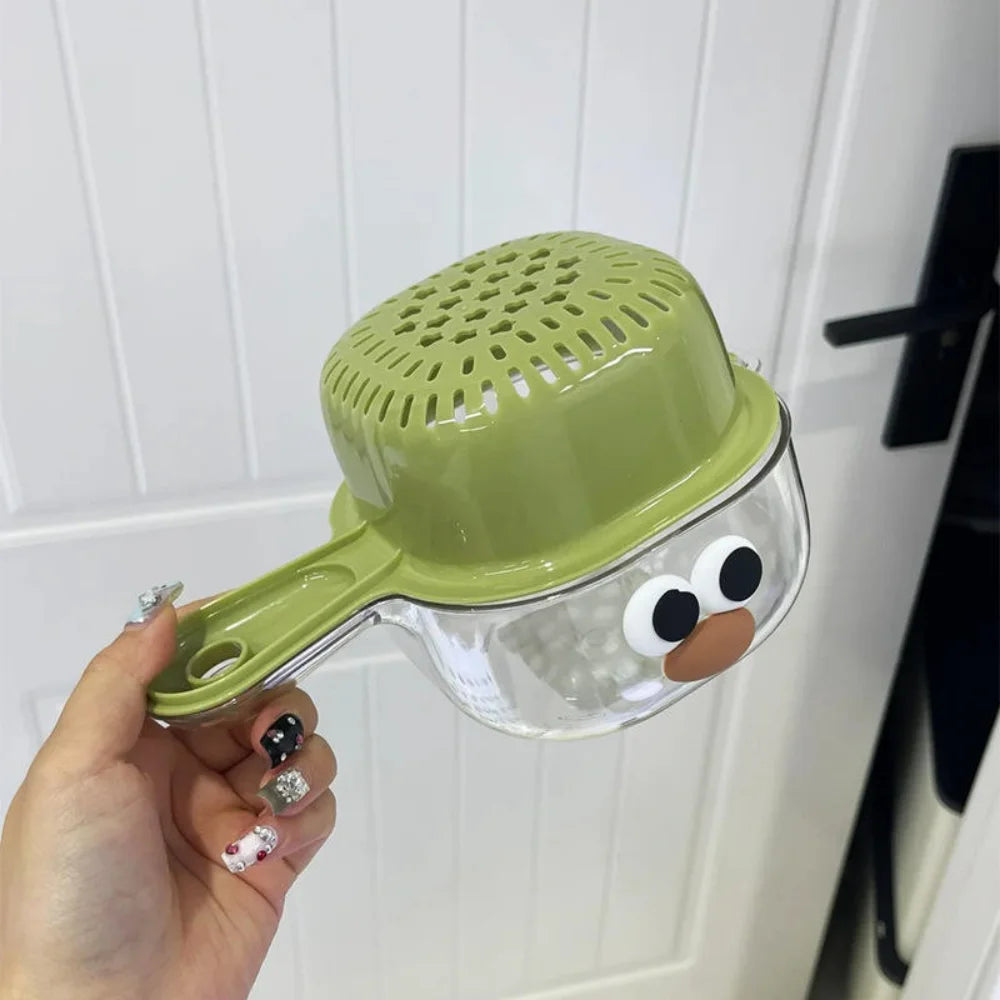 A hand holds a clear, double-layer wall-mounted fruit washing basket with a light green dome and a brown face design.