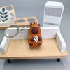 An adorable brown capybara charm with a white detail is displayed on a wooden tray next to a white electronic device.