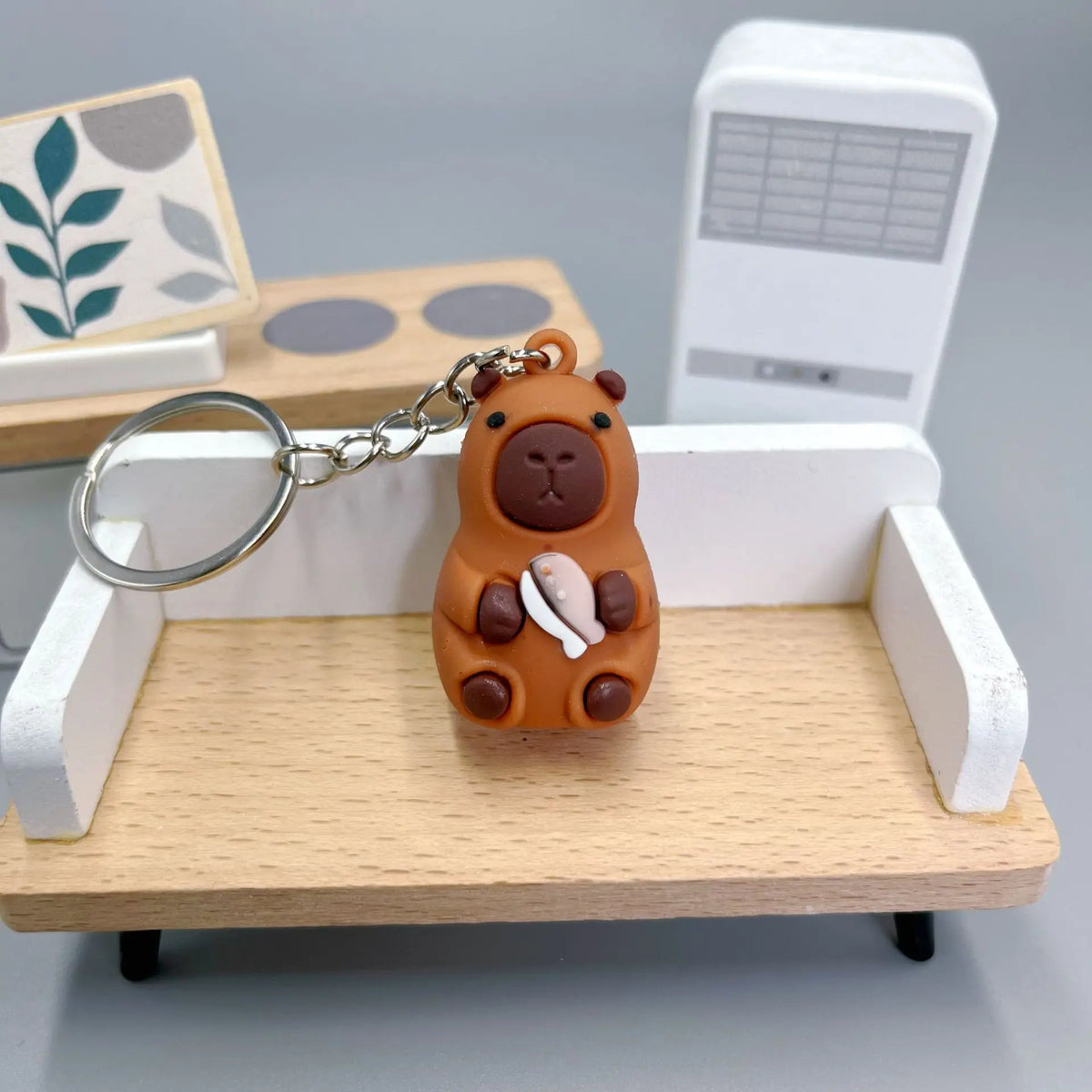An adorable brown capybara charm with a white detail is displayed on a wooden tray next to a white electronic device.
