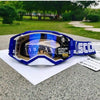 Blue and black motocross goggles resting on a gray surface with a blurred green outdoor background.