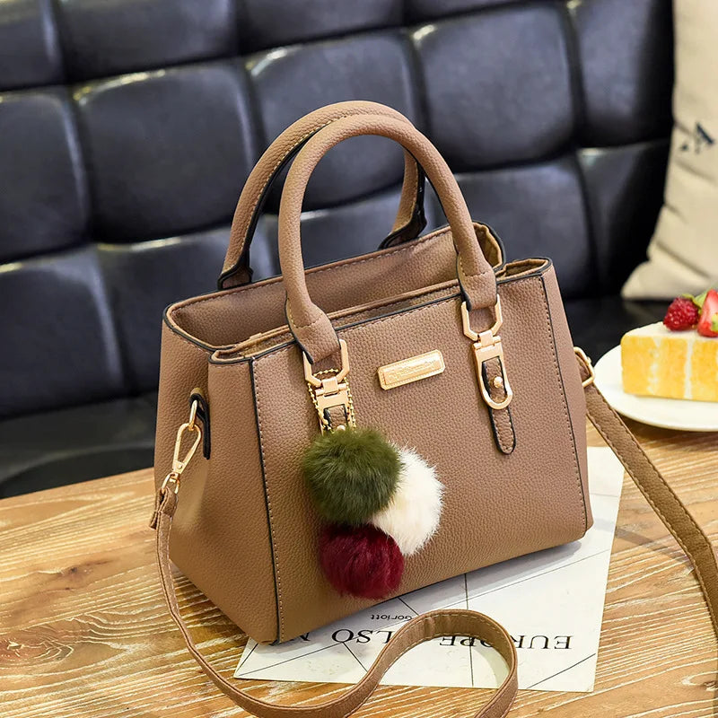 A tan PU leather shoulder bag with a tassel detail and pom-pom decoration resting on a wooden surface.
